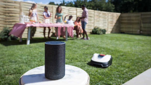 Amazon Alexa mit Miimo auf dem Rasen, Nahaufnahme, im Hintergrund eine Gartenparty.