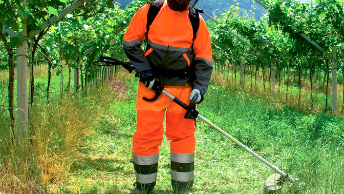 Model mit rückentragbarem Freischneider auf einem Feld.