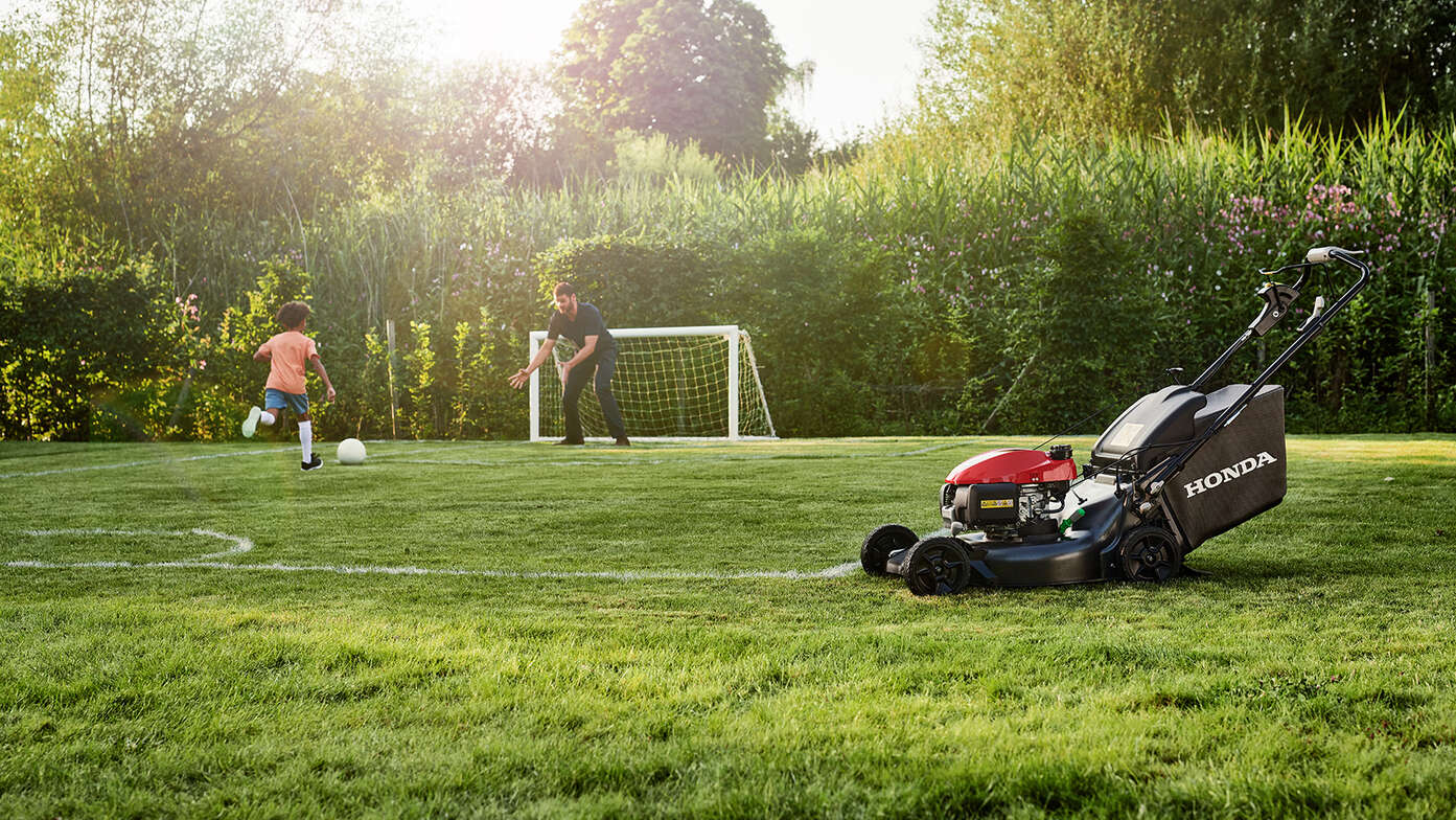 Mann und Kind beim Fußballspielen neben einem Honda HRN im Garten, Seitenansicht. 