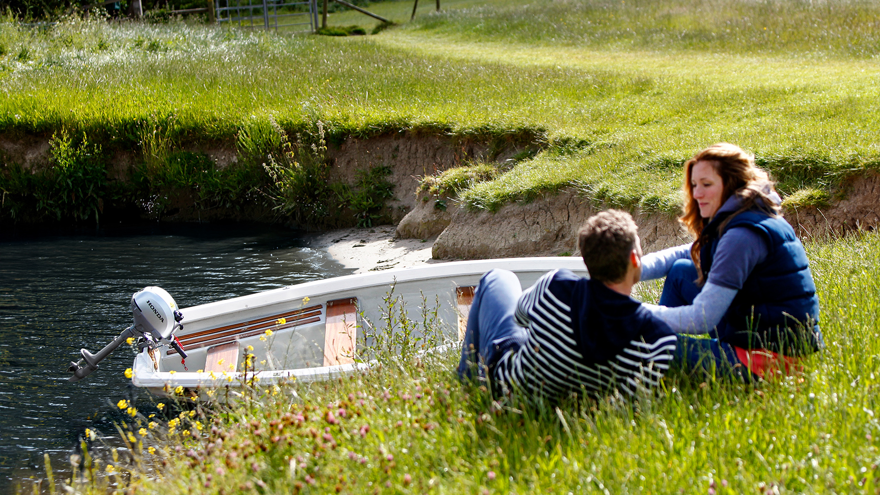 Zwei Personen, die neben einem im Wasser liegenden Boot am Ufer sitzen.