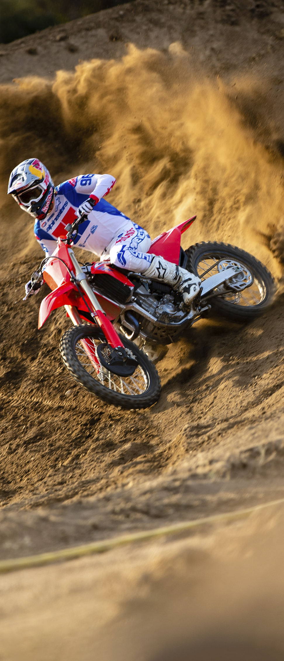 Ein Motocross-Fahrer in weißer, blauer und roter Rennkleidung legt sein rotes Motorrad in eine steile, sandige Böschung und wirbelt dabei eine große Welle Erde auf, während er kraftvoll durch die Kurve fährt.