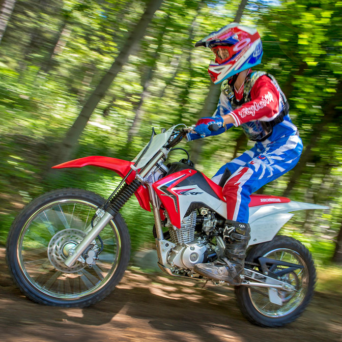 Seitenansicht der CRF125R mit Fahrer auf Geländefahrt bei einem Waldrennen. Nach links weisend.