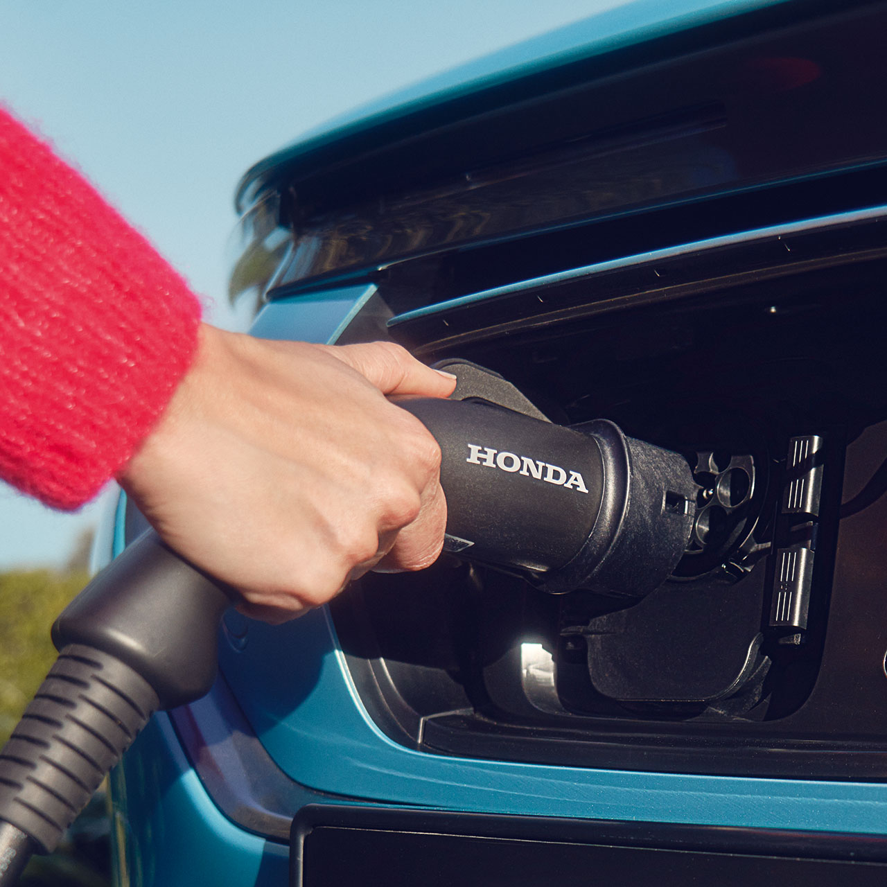 Eine Hand in einem roten Ärmel steckt ein Ladekabel für Honda-Elektrofahrzeuge in den Ladeanschluss eines blaugrünen Autos.