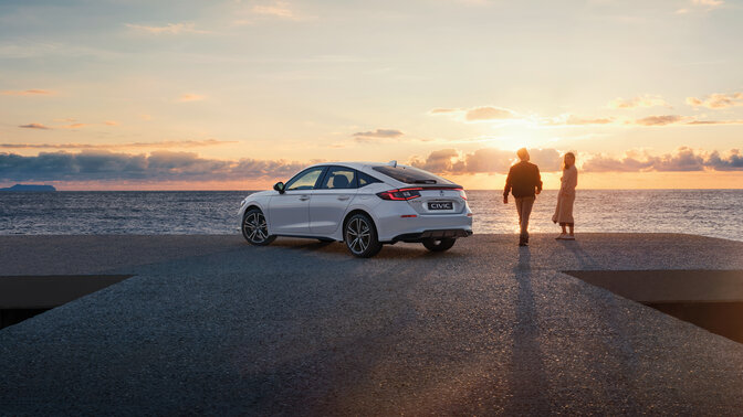 Honda Civic steht auf Straße am Meer bei Sonnenuntergang, daneben ein Paar.