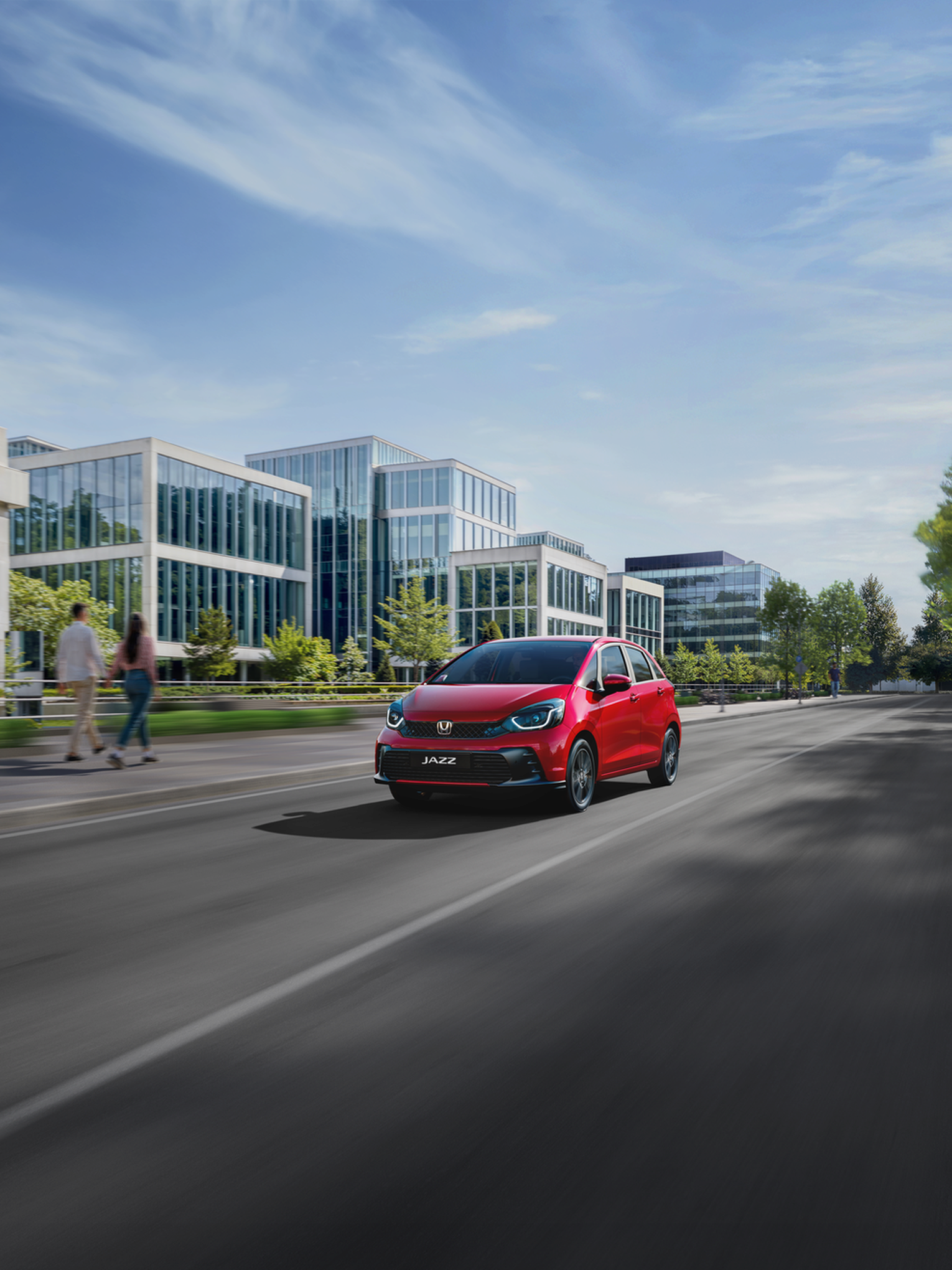 Honda Jazz in rot fährt eine Straße in einer Stadt entlang. Firmengebäude auf der linken Seite. Spaziergänger auf dem Fußweg