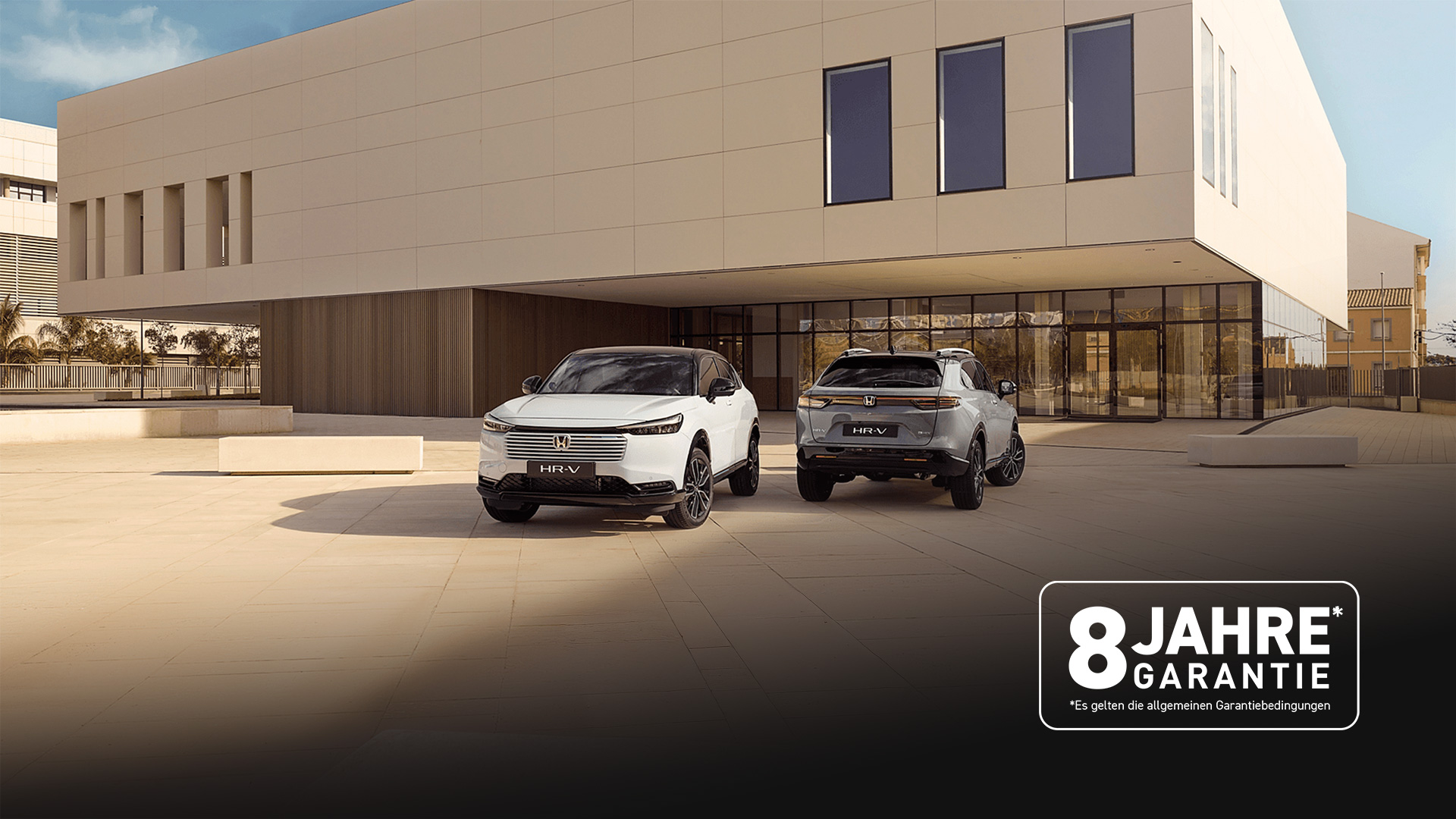 Zwei Honda HR-V SUVs, die vor einem modernen Gebäude geparkt sind, aus der Vorder- und Rückansicht – einer in Weiß, der nach vorne zeigt, und einer in Silber, der mit der Rückseite zum Betrachter zeigt.