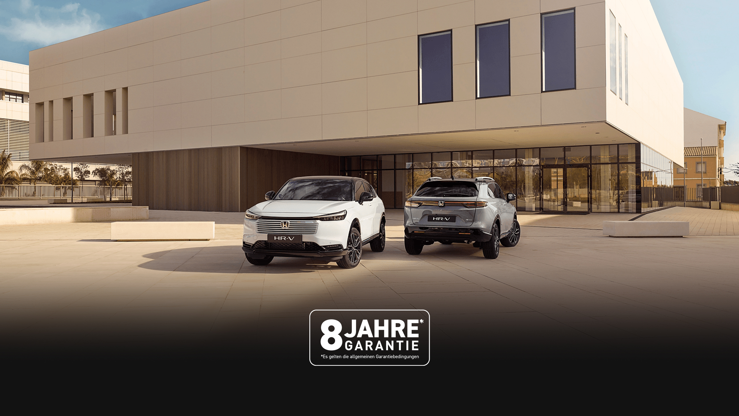 Zwei Honda HR-V SUVs, die vor einem modernen Gebäude geparkt sind, aus der Vorder- und Rückansicht – einer in Weiß, der nach vorne zeigt, und einer in Silber, der mit der Rückseite zum Betrachter zeigt.