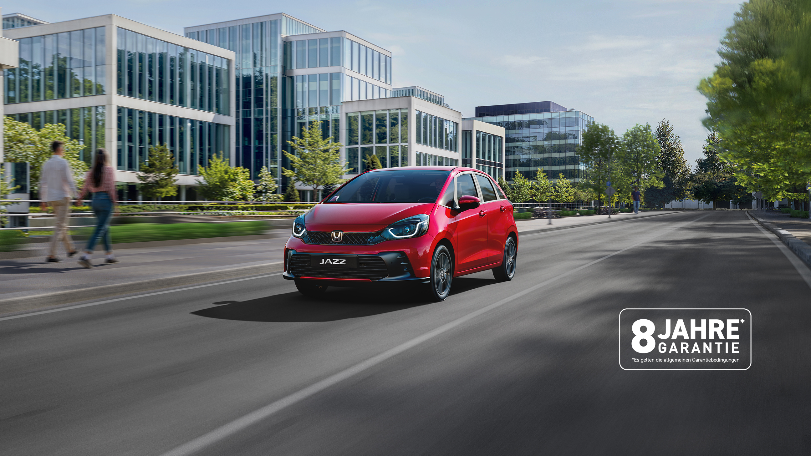 Honda Jazz in rot fährt eine Straße in einer Stadt entlang. Firmengebäude auf der linken Seite. Spaziergänger auf dem Fußweg