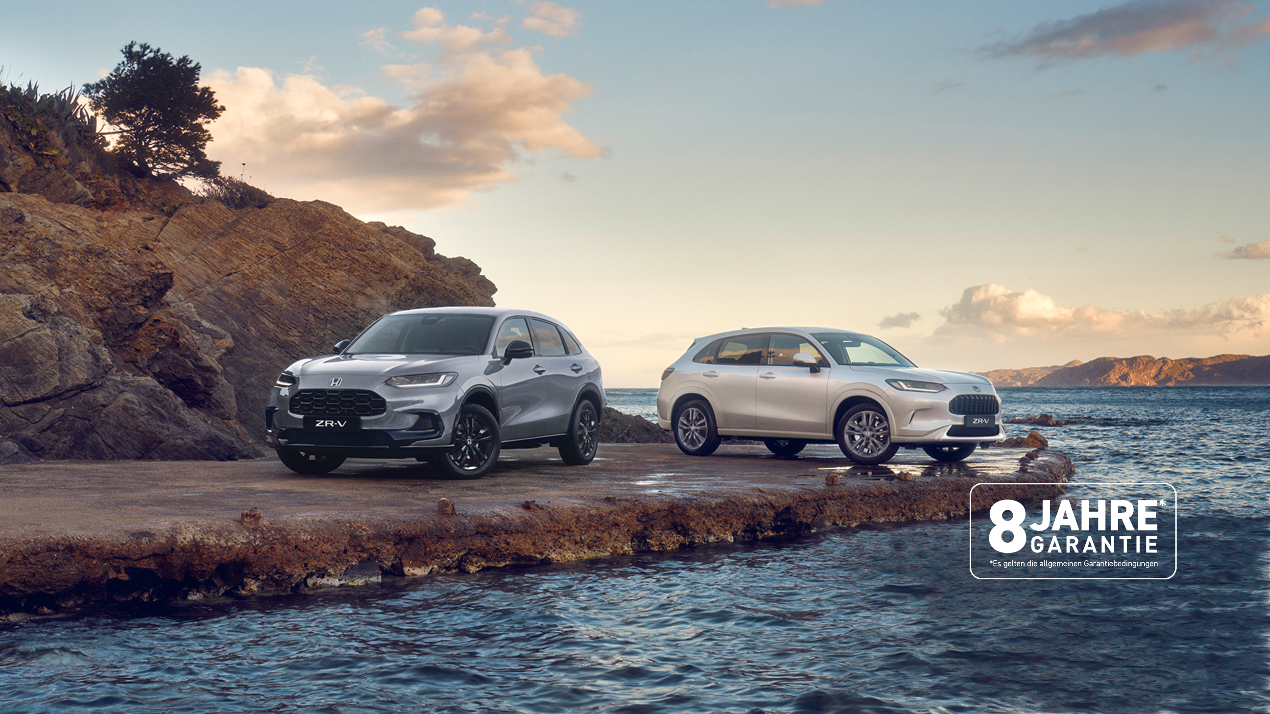 Zwei Honda ZR-V SUVs, one blue and one white, parken auf einer gebirgigen Küste, umgeben von Wasser im Vordergrund, und Gebirge im Hintergrund. Das Garantielogo über dem Auto