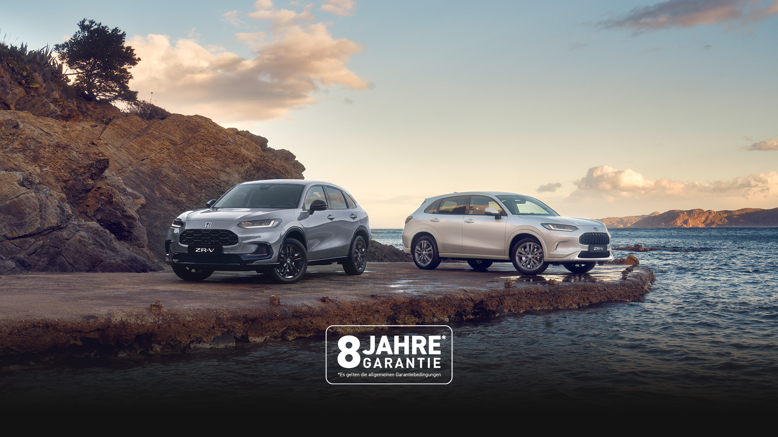 Two Honda ZR-V SUVs, one blue and one white, parked on a rocky coastal outcrop with water and hills in the background under a partly cloudy sky.