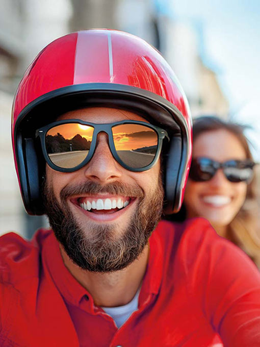 A bearded man and a woman enjoy a sunny ride on a motorbike in the city, both wearing helmets and sunglasses, radiating joy and friendship on a clear day.