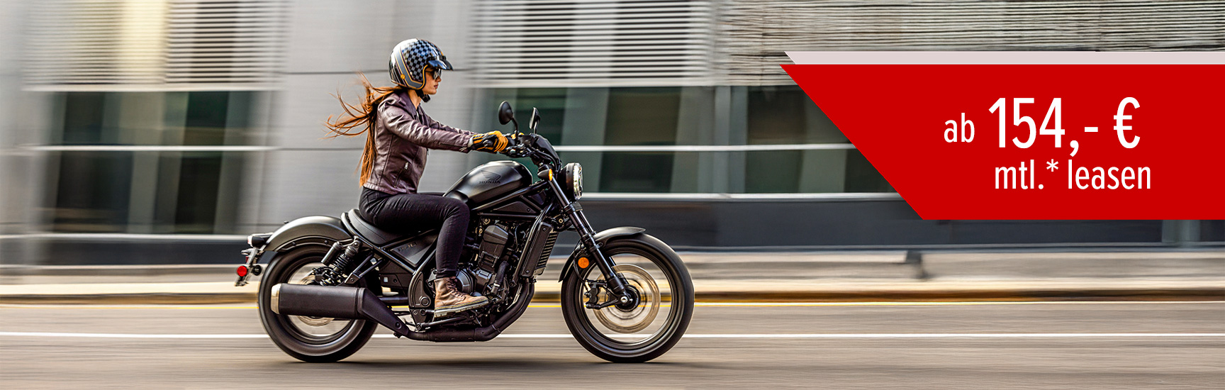 Eine Frau fährt auf einer Honda CMX1100 Rebel an einer Häuserfront vorbei.